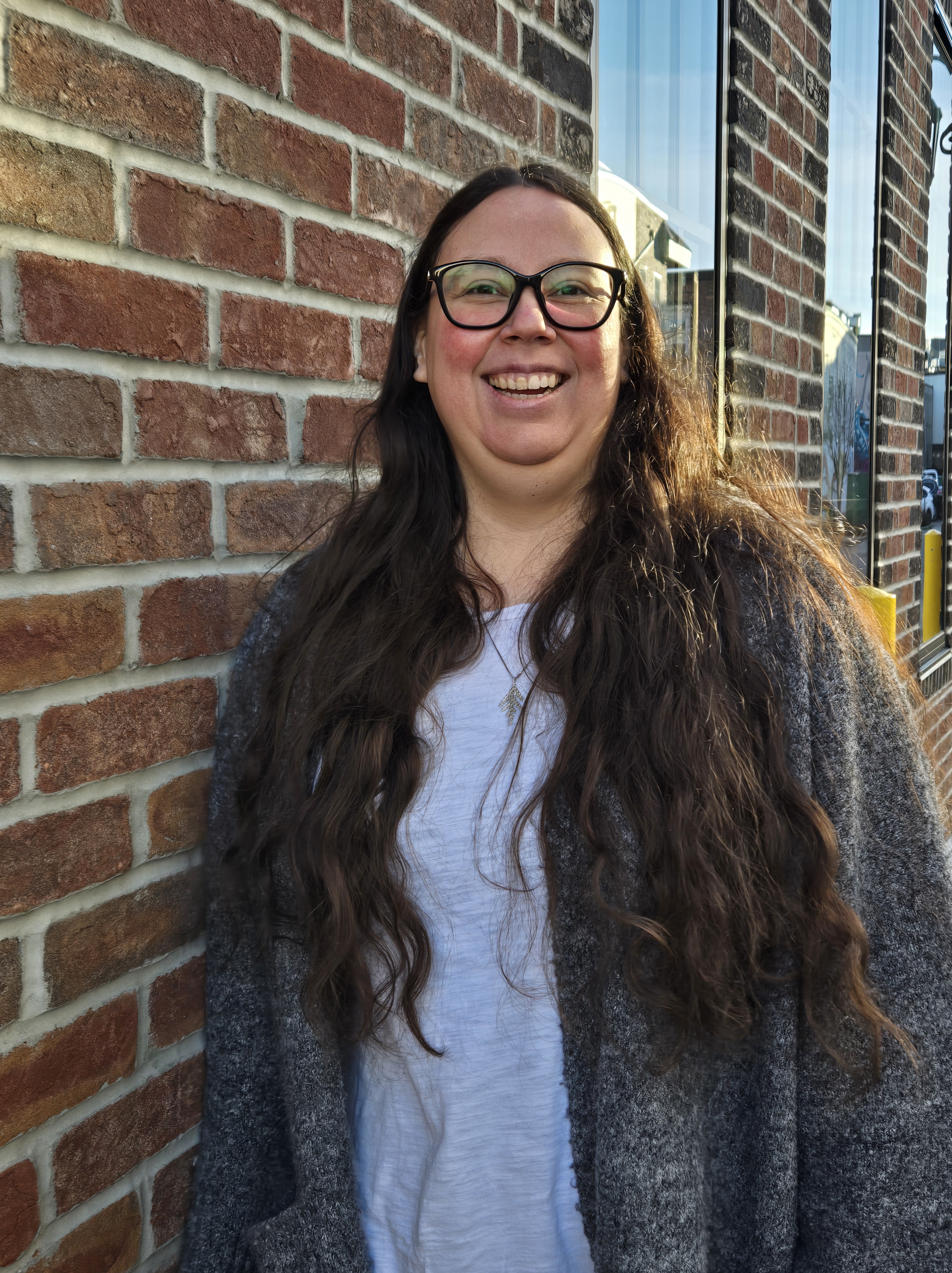 Woman with glasses and long hair wearing gray cardigan and white shirt leans on red brick wall.